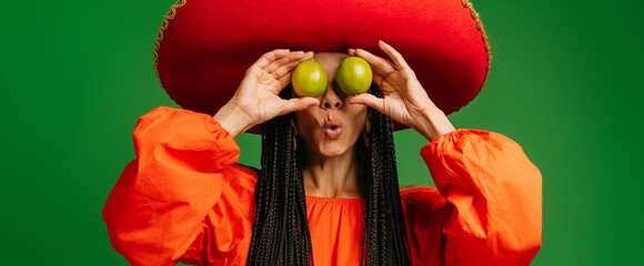 Surprised young Mexican woman in Sombrero covering eyes with fresh limes against green background - 749418346