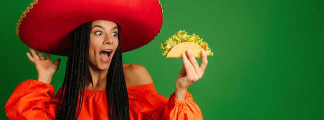 Attractive young Mexican woman in Sombrero holding taco and looking excited against green background - 749419535