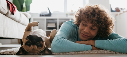 Handsome young curly man looking at his cute dog sleeping while lying on the floor at home together - 749422731