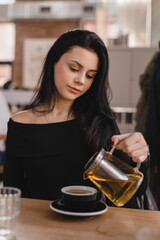 Brunette woman sitting at table drinking tea in restaurant. Teen woman wear black long sleeves top pouring green tea from glass teapot into cups in cafe. Happy girl relaxing and rest with friends.