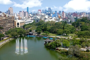 Fototapeta na wymiar Aerial scenery of Downtown Taichung, a vibrant metropolis in central Taiwan, with modern skyscrapers in background & a rainbow fountain in a pond surrounded by green forests in a park under sunny sky