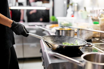 A chef in black attire sautés green vegetables in a bustling kitchen. Stainless steel pots and pans surround the stove. 