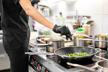A chef in black attire sautés green vegetables in a bustling kitchen. Stainless steel pots and...