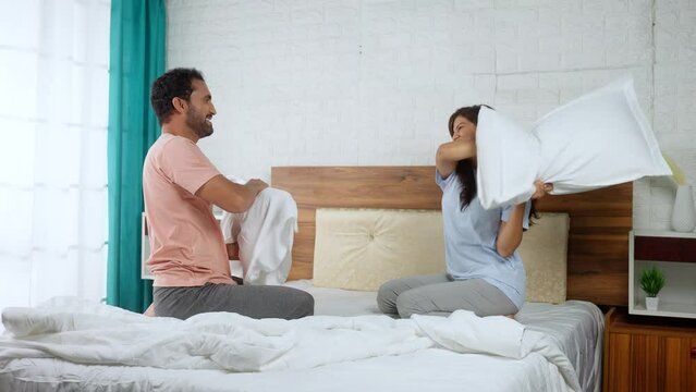 joyful Indian couples playing by fighting with pillow on bed - concept of relationship affection, happiness and romantic teasing
