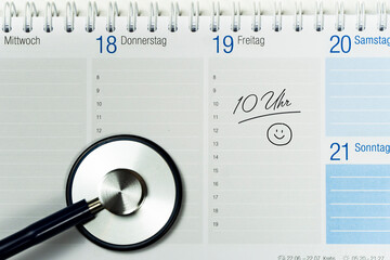 Teilansicht eines Tischkalenders, auf dem ein Termin notiert ist und auf dem das Ende eines Stethoskops zu sehen ist, horizontal
