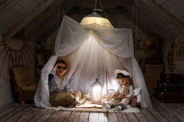 Children play in the attic of their house. Imagine themselves as tourists, explorers. Look through a telescope, study a world map, play in a makeshift tourist tent. Dreaming of travel and adventure. - 749453948