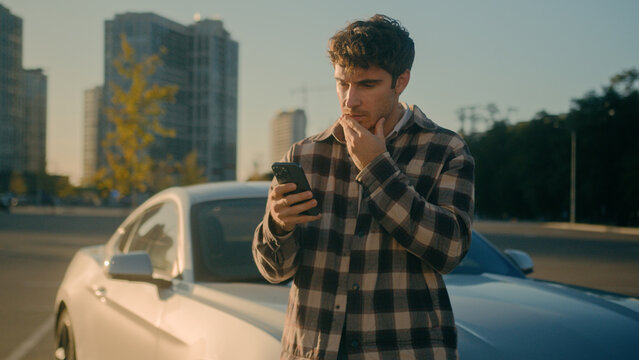 Caucasian confused man disappointed 30s guy millennial male businessman worried stressed car driver near luxurious modern auto on city parking outdoors reading bad news mobile phone problem loss upset