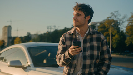 Successful Caucasian man stand near luxurious car in sunlight sunbeam outdoors happy male businessman hold smartphone browsing mobile phone internet buying new auto modern automobile dream in city