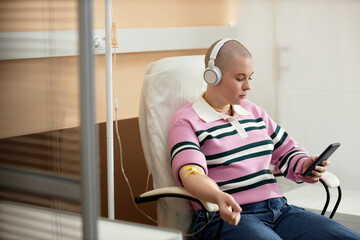 Portrait of bald young woman receiving IV treatment in clinic and using smartphone with headphones...