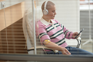 Side view portrait of bald young woman receiving IV treatment in clinic and using smartphone behind...