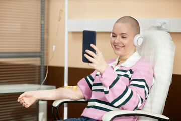Portrait of smiling bald woman receiving IV treatment in clinic and talking via video chat copy...