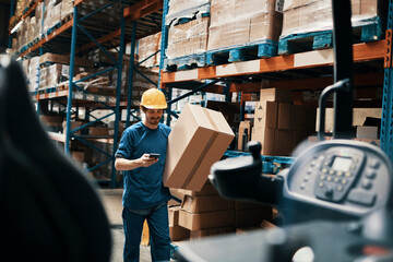 Young male warehouse worker using smartphone and carrying a box