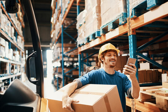 Young male warehouse worker using smartphone and carrying a box