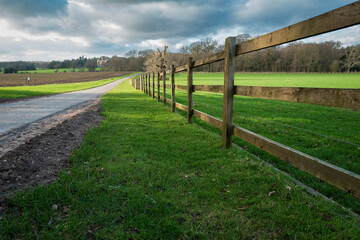 Empty rural road in the UK. Seen next to a long wooden fence adjacent to arable fields in the rural UK. The road leads to a distant English stately home seen in the distance.