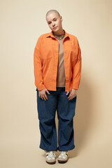 Vertical full length portrait of bald young woman wearing bright orange jacket and baggy jeans...