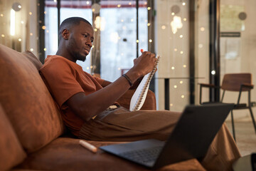 Side view portrait of Black young man drawing sketches sitting on couch at office or coworking space with low lighting copy space