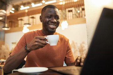 Front view portrait of smiling African American man using laptop in coffee shop and holding cup of...