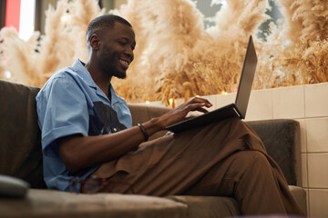 Side view portrait of cheerful African American man using laptop sitting on couch in office lounge and networking online