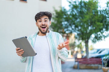 Handsome Arab man holding a tablet at outdoors with shocked facial expression