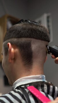 A barber man with a tattoo on his arm makes a haircut to a brunette guy in a barbershop, cuts his hair with a clipper close-up