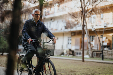Mature man with glasses and casual attire riding a bicycle with a basket through a serene city park, embracing an active lifestyle.