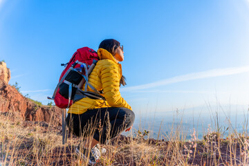 A young woman in the mountains, wearing a backpack and yellow jacket, enjoys hiking, admiring the...