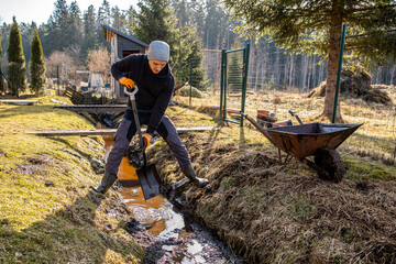 Man in workwear clearing a waterlogged garden path in early spring with a shovel and wheelbarrow amidst a rural landscape. © True Pixel Art