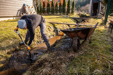 Man in protective gear tirelessly working to dredge a muddy trench with a shovel and wheelbarrow in a backyard during early spring.