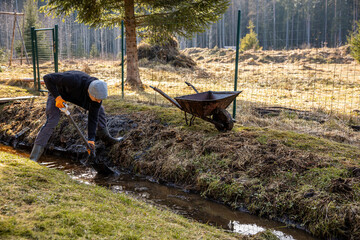 Man in workwear clearing a waterlogged garden path in early spring with a shovel and wheelbarrow...