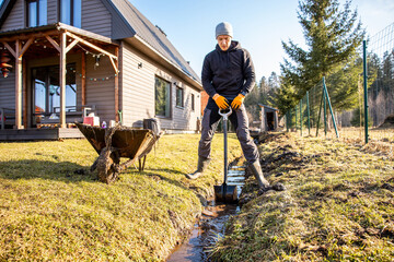 Determined man shoveling mud from a trench in his backyard on a sunny day, with a wheelbarrow and...