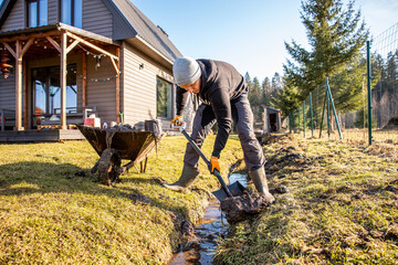 Determined man shoveling mud from a trench in his backyard on a sunny day, with a wheelbarrow and rural home in the background.