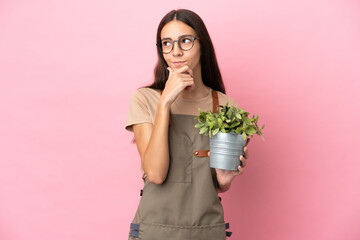Young gardener girl holding a plant isolated on pink background having doubts