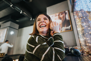 A happy young woman with braces is laughing heartily indoors with a bokeh of warm festive lights in the background, creating a joyful, relaxed atmosphere. - obrazy, fototapety, plakaty