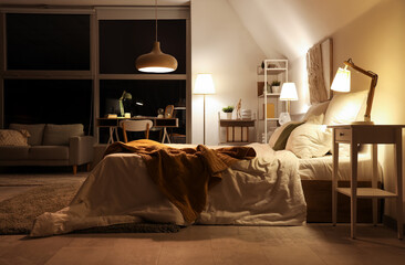 Interior of bedroom with plaid on bed, workplace and glowing lamps at night