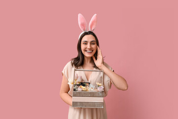 Beautiful young woman in bunny ears holding Easter basket with decorative cosmetics on pink background - obrazy, fototapety, plakaty