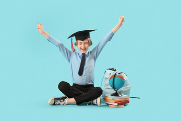 Little boy in graduation hat with books, globe and backpack on blue background. End of school year