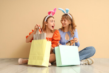 Surprised young women in bunny ears headbands unpacking shopping bags for Easter sitting near beige wall