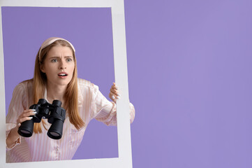 Young woman in pajamas with binoculars looking through window on lilac background