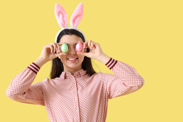 Beautiful young woman in bunny ears with Easter eggs on yellow background