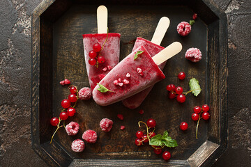 Homemade juicy red ice cream popsicles with fresh raspberries and red currant in wooden tray over brown concrete background. Berry sorbet. Top view. Healthy delicious summer dessert