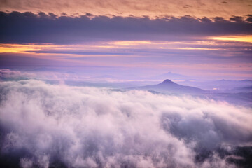 Tranquil sunrise with a lot of fog under orange-purple colorful sky, volcanic mountains in Central Bohemian Uplands (Czech: Ceske Stredohori). Impressive foggy morning landscape panorama. 