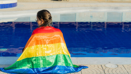 joven mujer latina sentada al lado de una piscina con la bandera del orgullo gay cubriendo su...