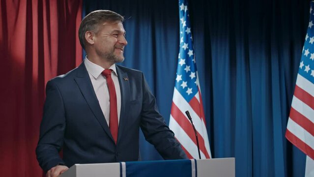 Medium lockdown shot of mature Caucasian male political candidate in formal suit, white shirt and tie, walking on stage, standing at tribune, smiling, waving, greeting public and nodding to opponent