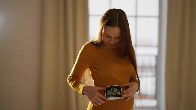 Pregnant woman looking ultrasound report. Pretty pregnant female watching her ultrasound report and touching her abdomen, admiring sonography picture of her unborn baby. Expecting baby concept.
