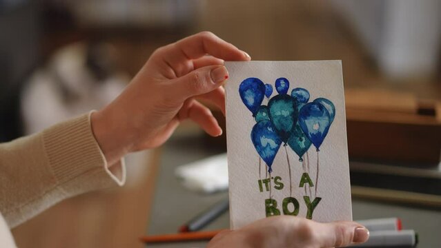 Slow motion. A woman holds a birthday card for her unborn child, which she drew with colored markers and watercolors, while sitting at her living room table. DIY greeting card