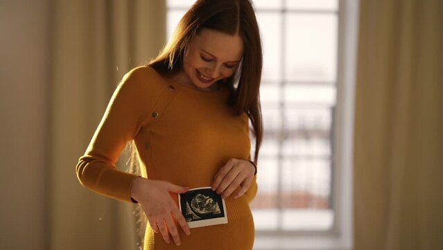 Pregnant woman looking ultrasound report. Happy pregnant female watching her ultrasound report and touching her abdomen, admiring sonography picture of her baby. Expecting baby loving concept.