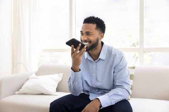Cheerful young African man dictating voice message on smartphone speaker, using virtual assistant, audio command service, smart home application on mobile phone, talking on speaker call