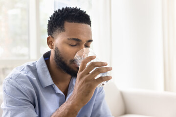 Calm thirsty young African man drinking fresh clear water with closed eyes from glass, keeping...