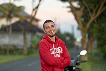 Portrait of a young man with a beard, smiling. A guy in a red sweatshirt is standing against the sunset.
