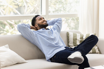 Happy carefree young African man relaxing on cozy home couch, stretching body on soft back with...